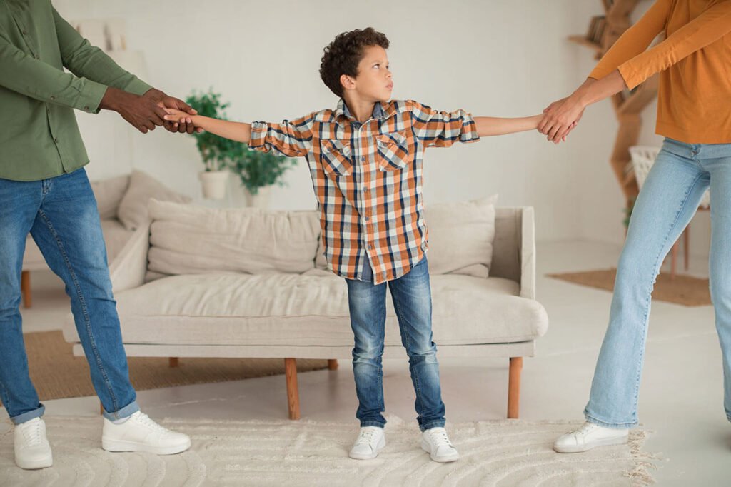 A young child with outstretched arms being held by two adults pulling in opposite directions in a domestic setting, illustrating a custody disagreement.